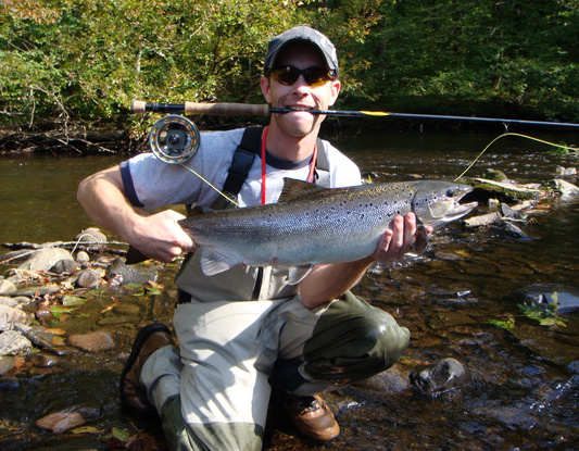 A photo of a fisherman holding a large fish while gripping his fishing pole in his mouth .